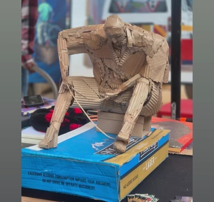 A cardboard sculpture of a seated, slouched figure built from layered cardboard strips, mounted on a carton box